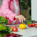 A person in a pink shirt reaching for a lemon surrounded by fresh vegetables.
