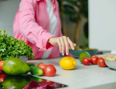 A person in a pink shirt reaching for a lemon surrounded by fresh vegetables.