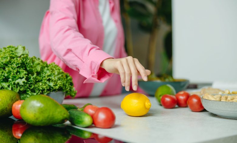 A person in a pink shirt reaching for a lemon surrounded by fresh vegetables.
