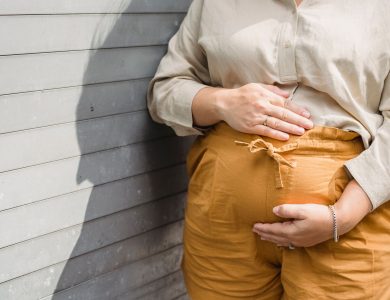 Crop unrecognizable pregnant female in shorts and shirt standing near wall and touching tummy