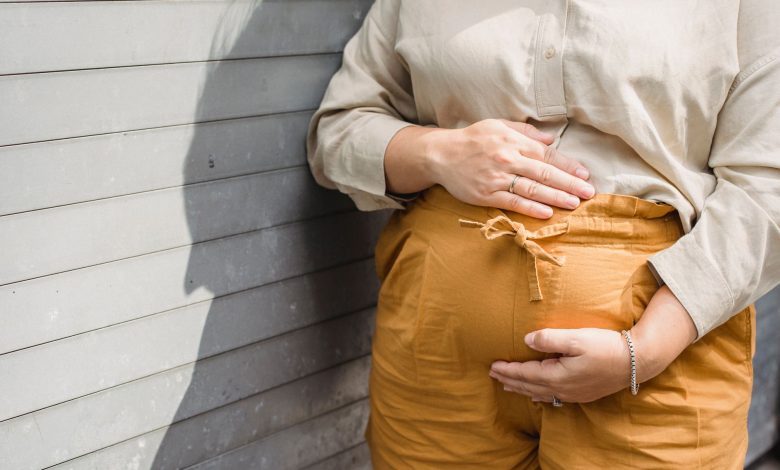 Crop unrecognizable pregnant female in shorts and shirt standing near wall and touching tummy