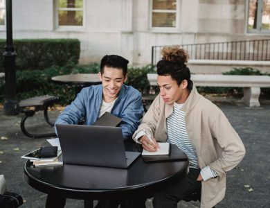 Smiling multiracial male classmates sitting at table and browsing netbook while taking notes in notebook
