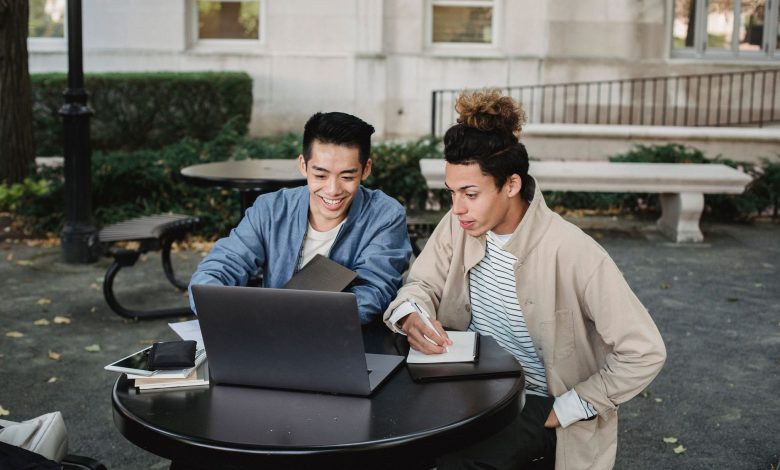 Smiling multiracial male classmates sitting at table and browsing netbook while taking notes in notebook
