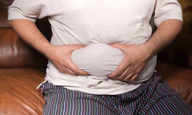A close-up view of a man holding his tummy while sitting on a couch, highlighting lifestyle choices.