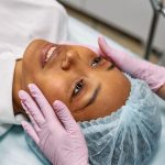 A woman receiving a facial treatment in a spa, wearing a hairnet while hands in gloves gently touch her face.