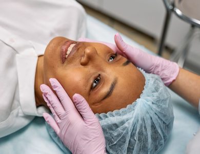 A woman receiving a facial treatment in a spa, wearing a hairnet while hands in gloves gently touch her face.
