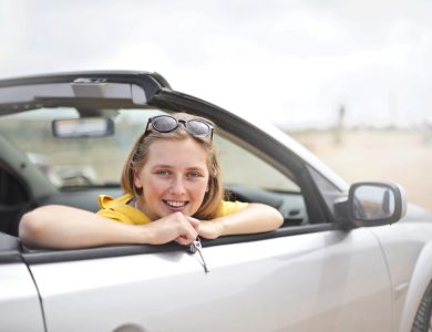 A young woman smiles joyfully while leaning out of a convertible car, enjoying a sunny day.